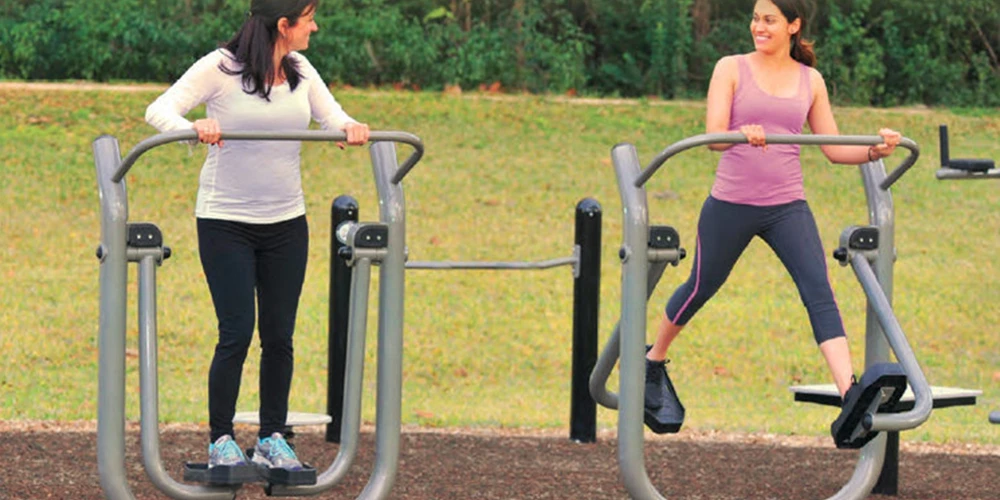 People using outdoor cardio fitness machines like air walkers and ellipticals in a park gym setting